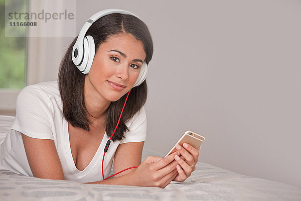 Hispanische Frau liegt auf dem Bett und hört mit Kopfhörern Handy
