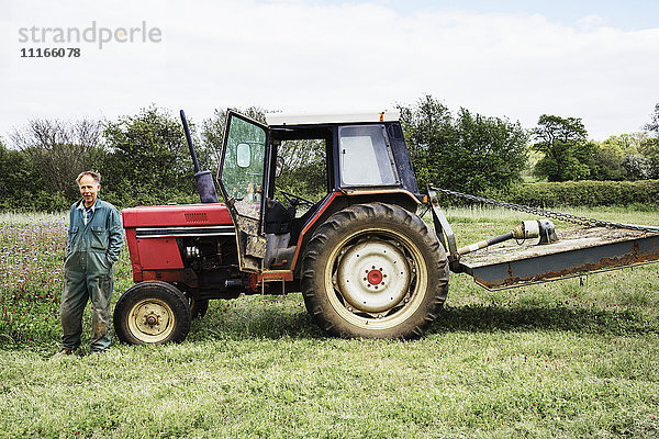 Ein stehender roter Traktor und ein an die Motorhaube gelehnter Fahrer auf einem Feld im Sommer.