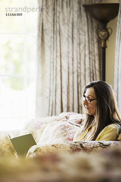 Ältere Frau mit langen braunen Haaren  die auf einem Sofa sitzt  ein Buch liest und eine Lesebrille trägt.