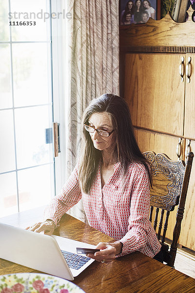Ältere Frau mit langen braunen Haaren  die an einem Tisch sitzt  einen Laptop-Computer benutzt und eine Lesebrille trägt.