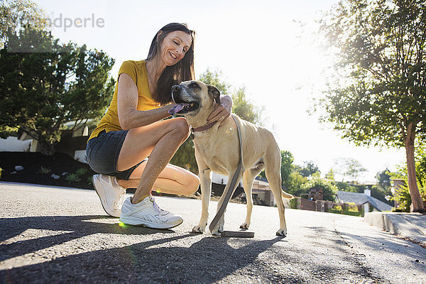 Ältere Frau in Shorts  kniend auf der Straße  einen Hund streichelnd.