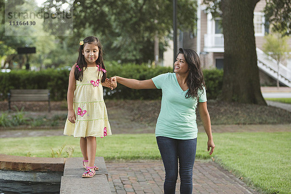 Hispanische Mutter hält die Hand ihrer Tochter  die auf einer Parkmauer läuft