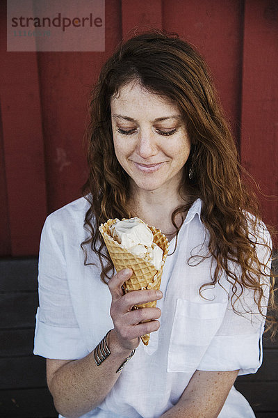 Frau mit langen braunen Haaren sitzt auf einer Bank und isst Eiscreme.