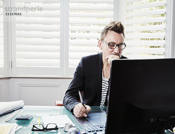 Ein Mann mit Brille sitzt an einem Schreibtisch in einem Büro und schaut auf einen Computerbildschirm.