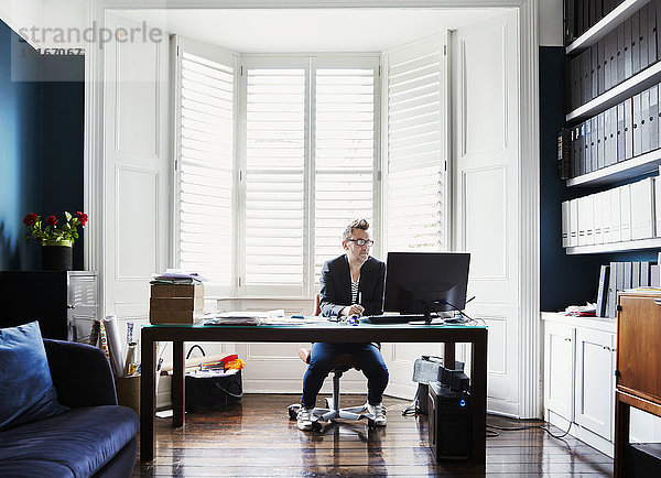Ein Mann in Anzug und Turnschuhen mit Brille  an einem Schreibtisch in einem Erkerfenster sitzend. Ein helles  luftiges Büro mit Akten auf Regalen.