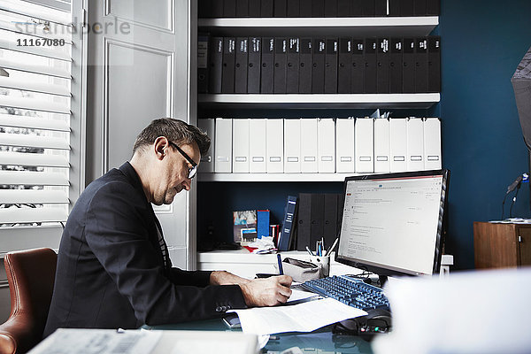 Ein Mann sitzt an einem Schreibtisch in einem Büro und schreibt auf einem Blatt Papier. Ordentliche Aktenreihen in den Regalen und Papiere auf dem Schreibtisch.