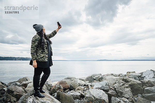 Mixed Race Frau steht auf Felsen am Meer posiert für Handy-Selfie