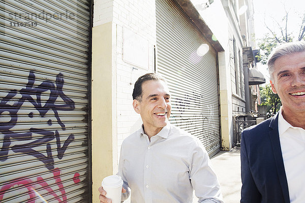 Zwei Männer gehen eine Straße entlang  vorbei an einem Stahlfensterladen an einem mit Graffiti bemalten Gebäude.