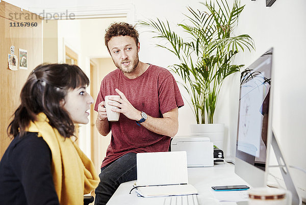 Eine Frau  die an einem Schreibtisch arbeitet  und ein Mann  der mit einer Kaffeetasse auf dem Schreibtisch sitzt  sich unterhält und auf den Computerbildschirm schaut.