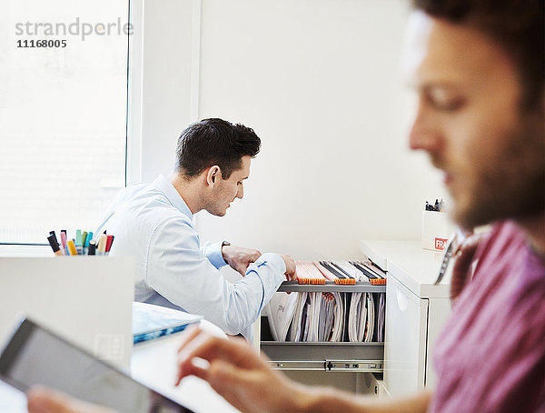 Ein Mann  der in einem Büro Akten durchsieht  und ein Mann  der sein digitales Tablett benutzt.