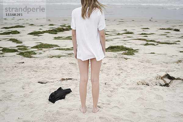 Rückansicht eines Mädchens mit blonden Haaren in einem weißen T-Shirt  das an einem Sandstrand steht.