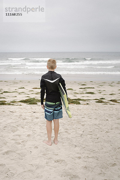 Blonder Junge  der an einem Sandstrand steht und ein Bodyboard hält.