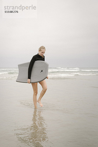 Blondes Mädchen  das an einem Sandstrand entlang geht und ein Bodyboard trägt.
