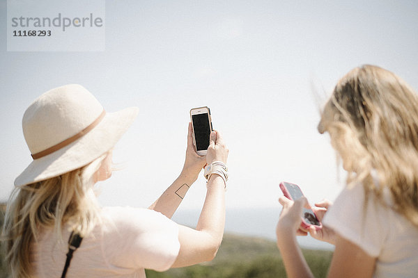 Frau und junges Mädchen mit langen blonden Haaren stehen im Freien  halten ein Mobiltelefon hoch und machen ein Foto.