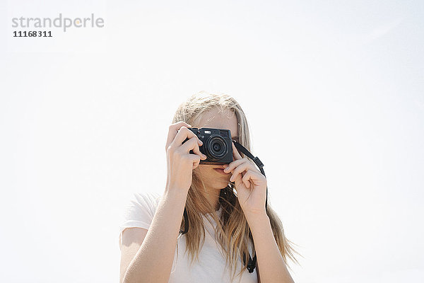 Teenager-Mädchen mit langen blonden Haaren  das durch eine Kamera schaut und ein Foto macht.