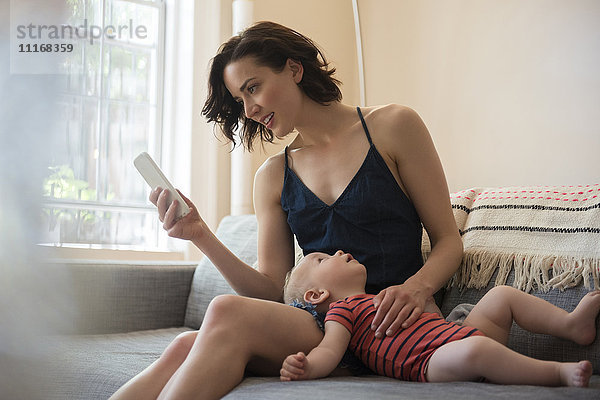Kaukasische Mutter sitzt mit ihrem kleinen Sohn auf dem Sofa und schreibt eine SMS mit dem Handy