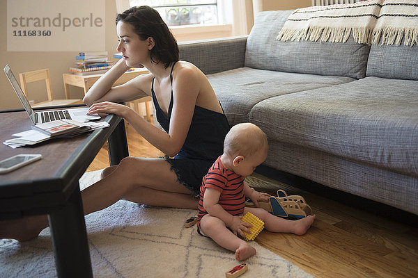 Kaukasische Mutter sitzt auf dem Boden und benutzt einen Laptop neben ihrem kleinen Sohn