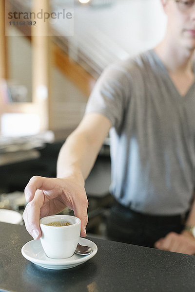 Mann  der an einem Tresen in einem Café steht und nach einer Tasse Espresso-Kaffee greift.