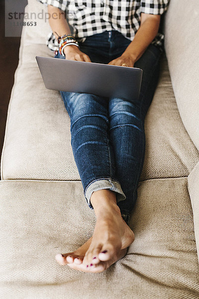 Frau in Jeans  die auf einem Sofa liegt und einen Laptop benutzt.