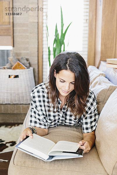Frau mit langen braunen Haaren liegt auf einem Sofa und liest ein Buch.