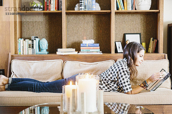 Frau mit langen braunen Haaren liegt auf einem Sofa und liest ein Buch.