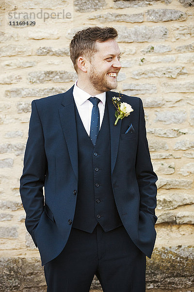 Ein Mann in einem Anzug mit einem Knopfloch  die Hände in den Taschen  lächelnd.