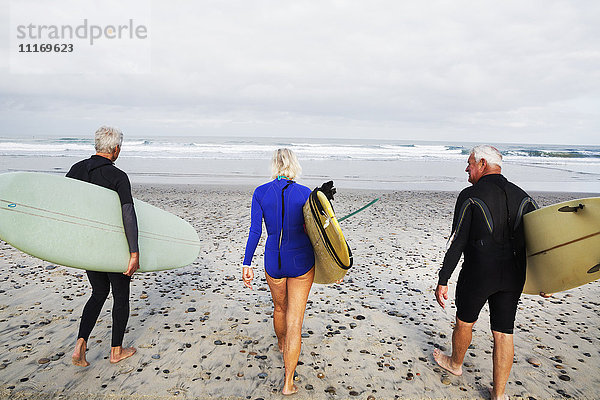 Ältere Frau und zwei ältere Männer an einem Strand  in Neoprenanzügen und mit Surfbrettern.