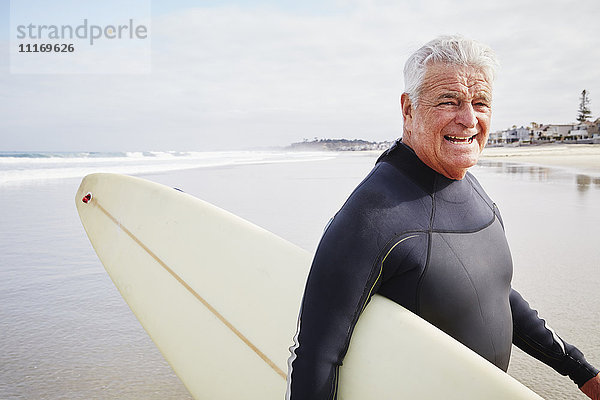Lächelnder älterer Mann steht am Strand  trägt einen Neoprenanzug und hat ein Surfbrett dabei.