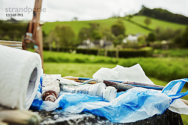 Eine Staffelei und eine Künstlerausrüstung auf einer Plastiktüte. Tuben mit Farbe und Papier  im Freien. Szenische Ansicht.