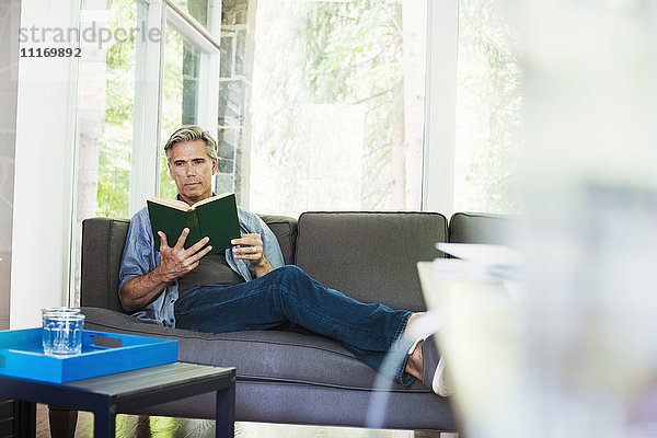 Ein Mann sitzt am Fenster auf einem Sofa und liest ein Buch.