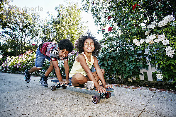 Gemischtrassiger Junge schiebt seine Schwester auf dem Skateboard über den Bürgersteig