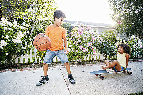 Gemischtrassiges Mädchen auf Skateboard  das seinem Bruder beim Basketballspielen zusieht