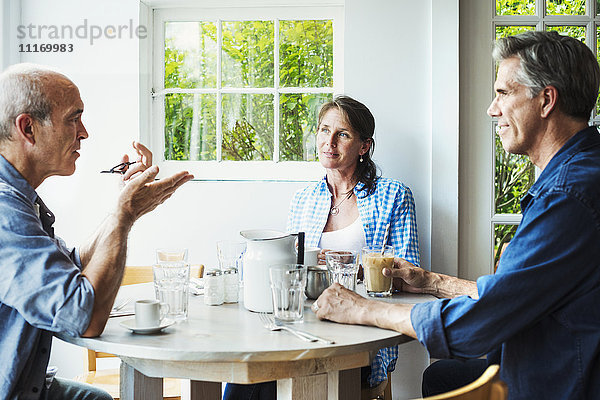Zwei Männer und eine Frau sitzen an einem Kaffeehaustisch und unterhalten sich.