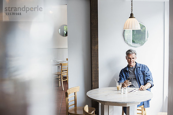Ein Mann sitzt an einem Coffeeshop-Tisch und schaut auf sein Smartphone. Unscharfer Vordergrund.