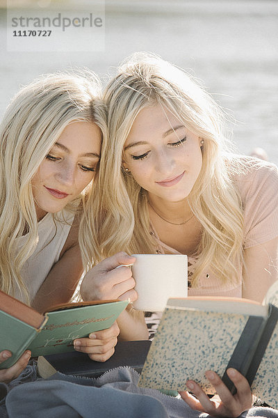 Zwei blonde Schwestern liegen auf einem Steg und lesen ein Buch.