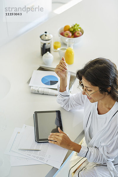 Frau im Bademantel bei der Arbeit am digitalen Tablet am Küchentisch