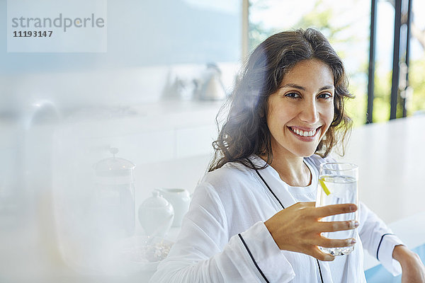 Porträt einer lächelnden Frau im Bademantel  die in der Küche Wasser trinkt