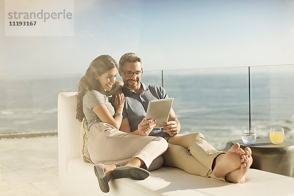 Paar entspannt mit digitalem Tablet auf sonnigem Luxusbalkon mit Meerblick