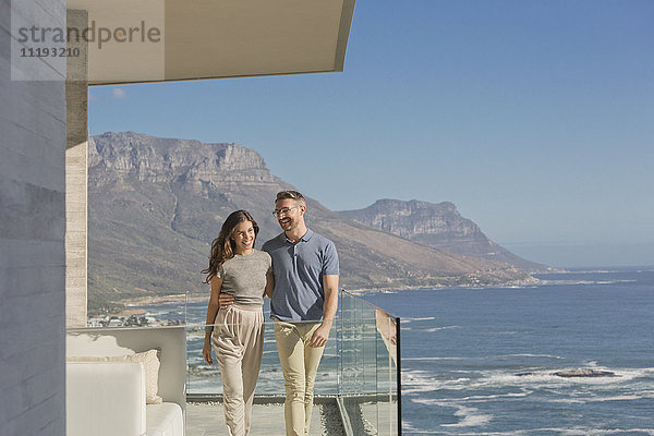 Lächelndes Paar  das auf einem luxuriösen Balkon mit sonnigem Meer- und Bergblick spazieren geht