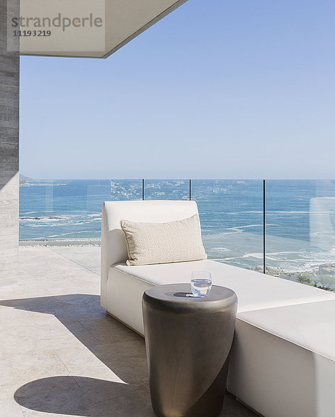 Chaise Lounge und Wasserglas auf sonnigem Luxusbalkon mit Meerblick