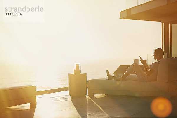 Silhouette Frau mit Handy auf Chaise Lounge auf Luxus-Balkon mit Sonnenuntergang Blick auf das Meer