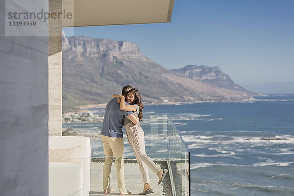 Paar  das sich auf dem sonnigen Luxusbalkon mit Blick auf das Meer und die Berge umarmt