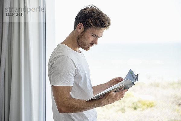 Junger Mann beim Lesen einer Zeitschrift am Glasfenster