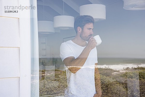 Junger Mann schaut durchs Fenster mit einer Tasse Kaffee.