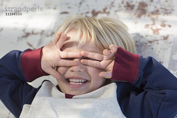 Verspielter kleiner Junge  der sich hinlegt und seine Augen mit den Händen bedeckt.