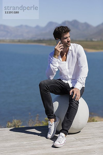 Mann sitzt auf einer Steinkugel und spricht auf einem Handy