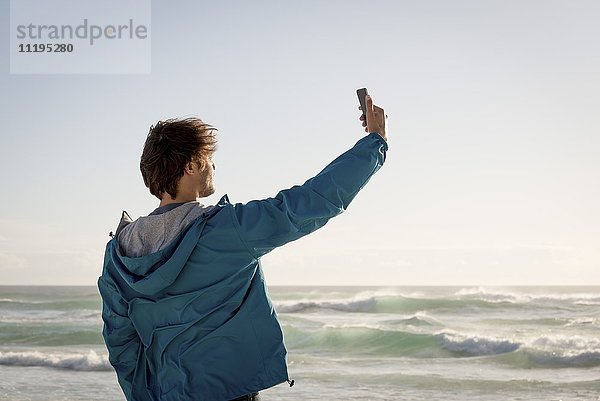 Glücklicher junger Mann nimmt Selfie mit Fotohandy am Strand