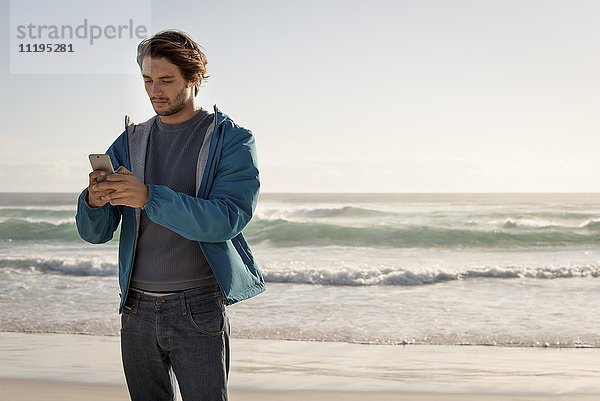 Glücklicher junger Mann mit dem Handy am Strand