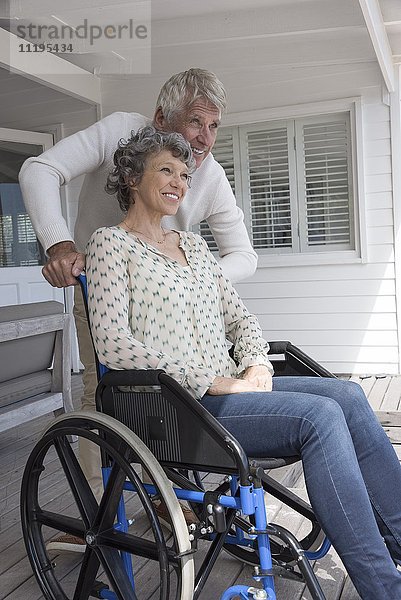 Glücklicher älterer Mann  der seiner Frau im Rollstuhl auf der Veranda hilft.