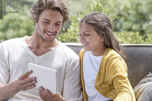 Glücklicher Vater mit seiner kleinen Tochter  die ein digitales Tablett auf einem Schaukelstuhl benutzt.